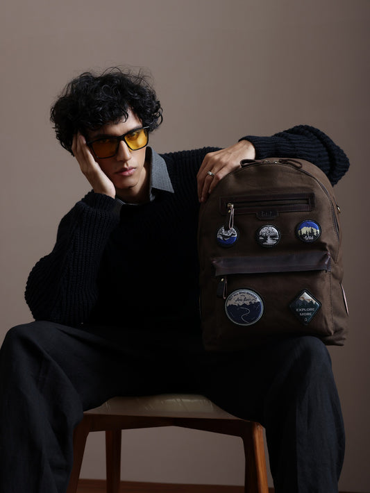 Man holding the Nicobar brown canvas and reclaimed leather travel backpack with embroidered adventure badges in an indoor lifestyle portrait.