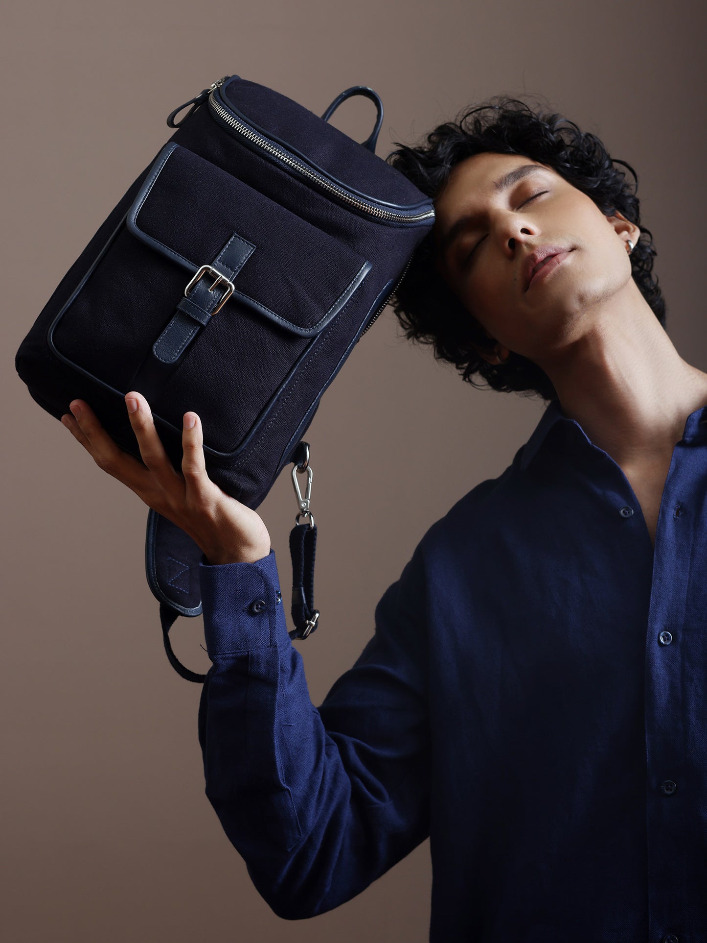 Man holding the Studio Ek Inch navy canvas and reclaimed leather mini crossbody backpack with front buckle pocket, top zip compartment, silver nickel hardware, and adjustable shoulder strap.