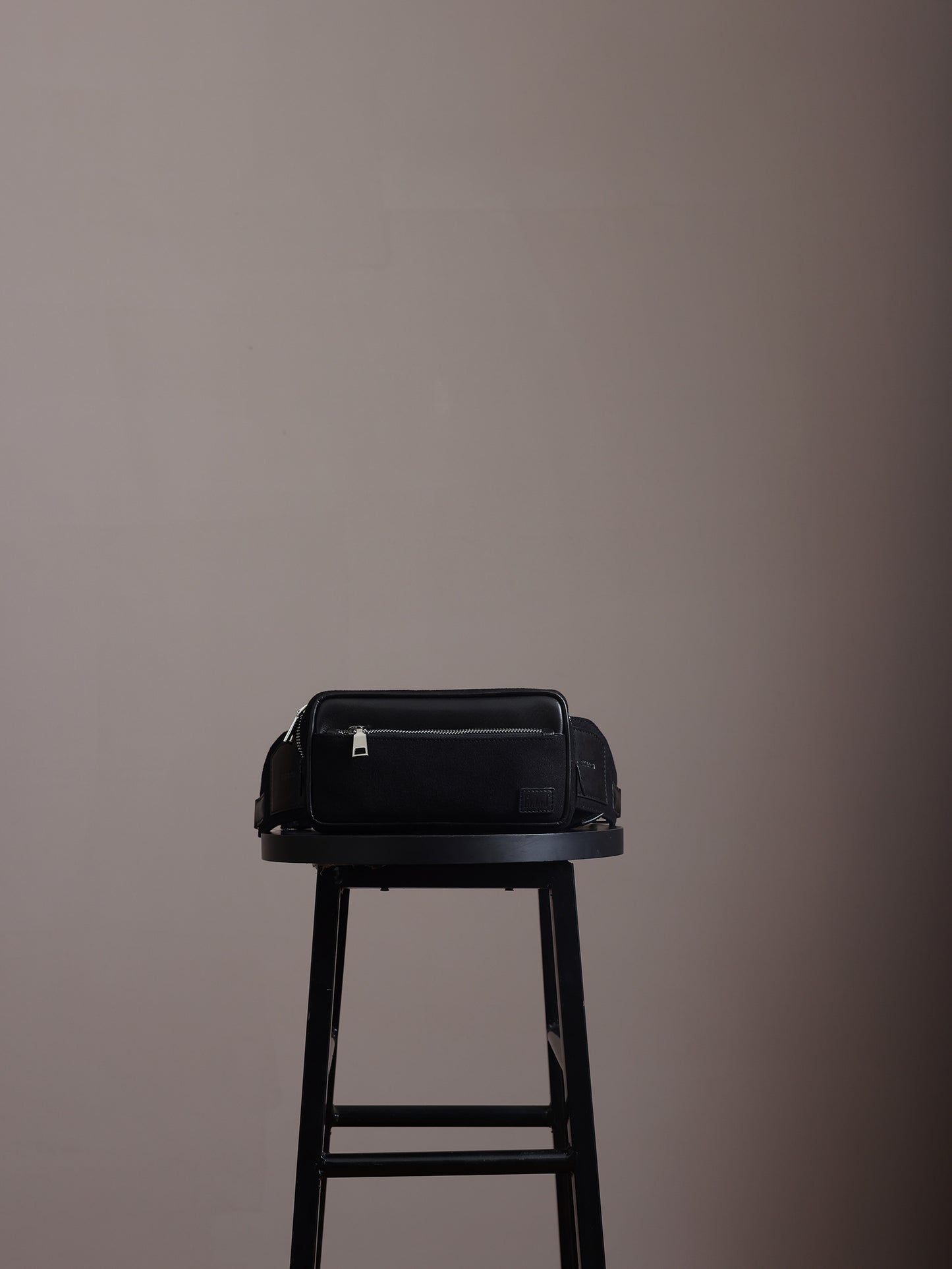 Minimal studio shot of the Studio Ek Inch Metropolitan crossbody bag in black waterproof canvas and reclaimed leather placed on a stool, featuring a front zip pocket and premium hardware. Designed as a premium crossbody bag for men, it blends contemporary aesthetics with functional storage for travel and everyday use.