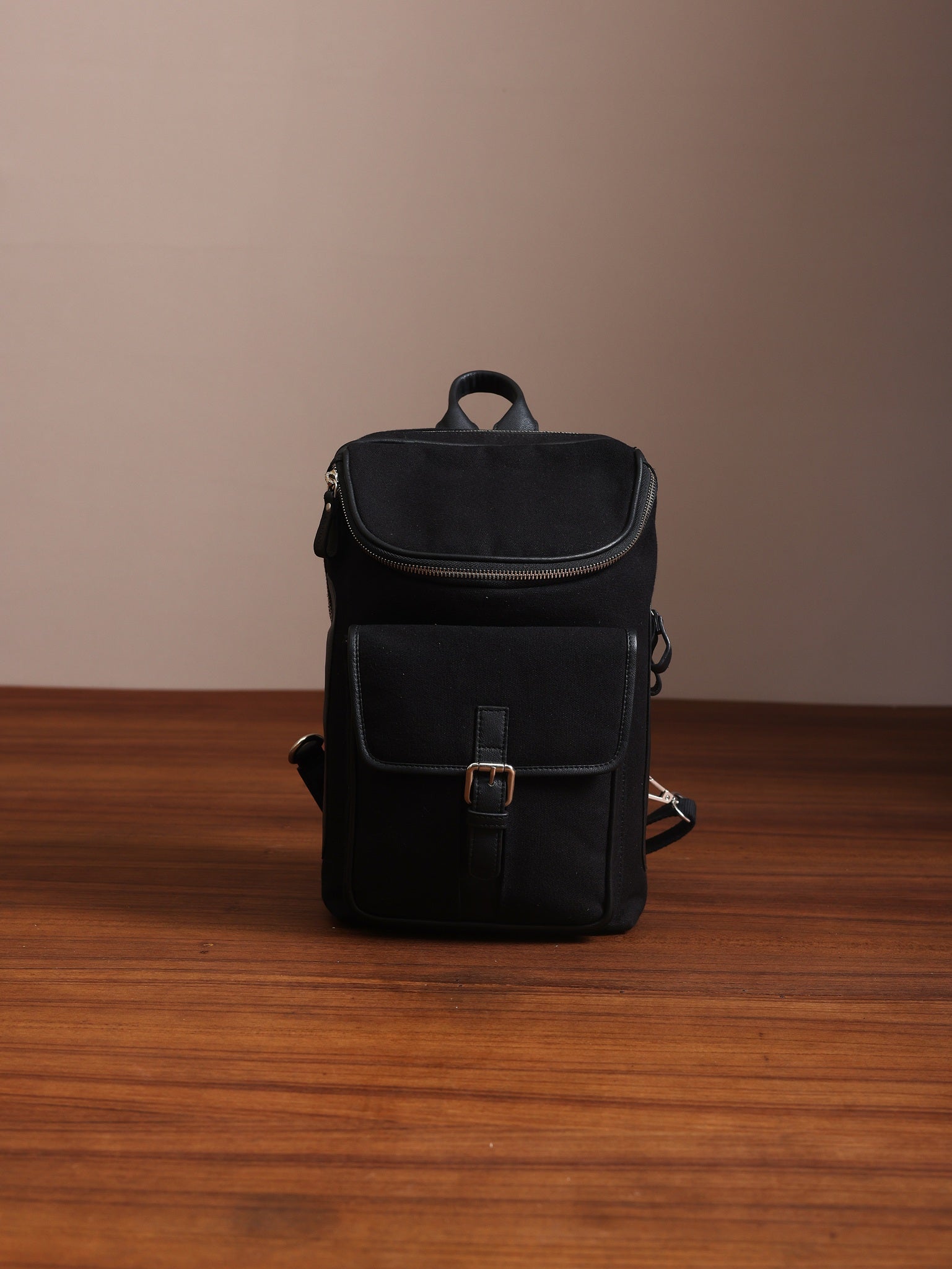 Studio Ek Inch black canvas and reclaimed leather mini crossbody backpack kept on wooden textured floor background.