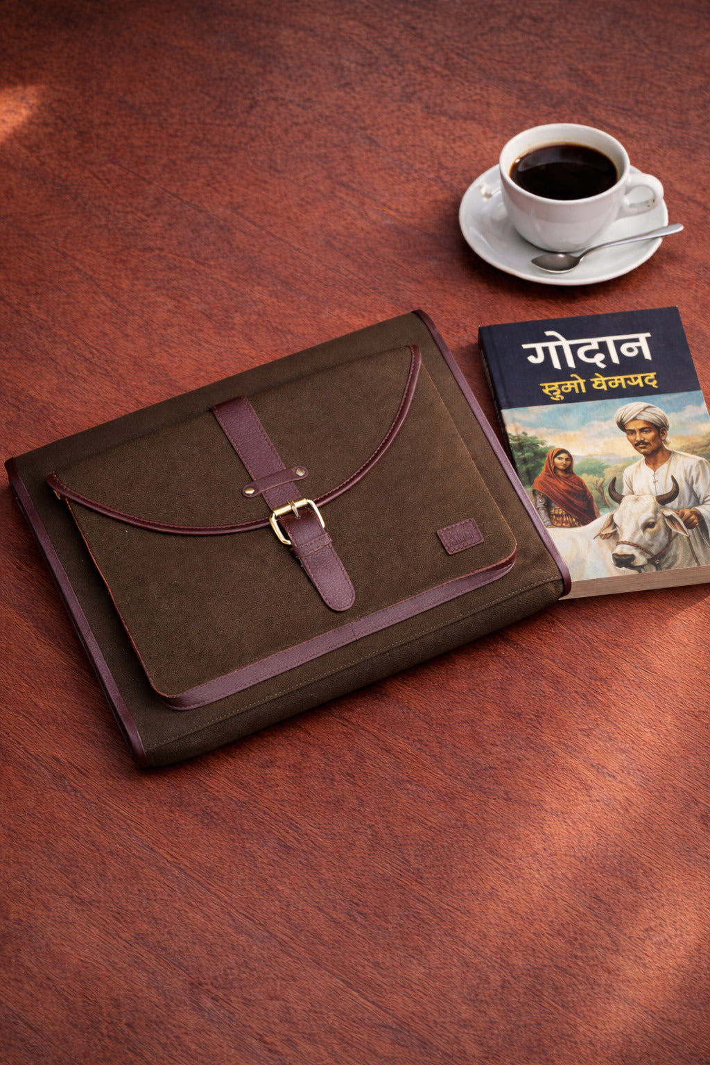 Leather and canvas Passage Sleeve laptop case with flap closure placed on wooden table beside coffee and book, designed for 16-inch laptop and iPad.