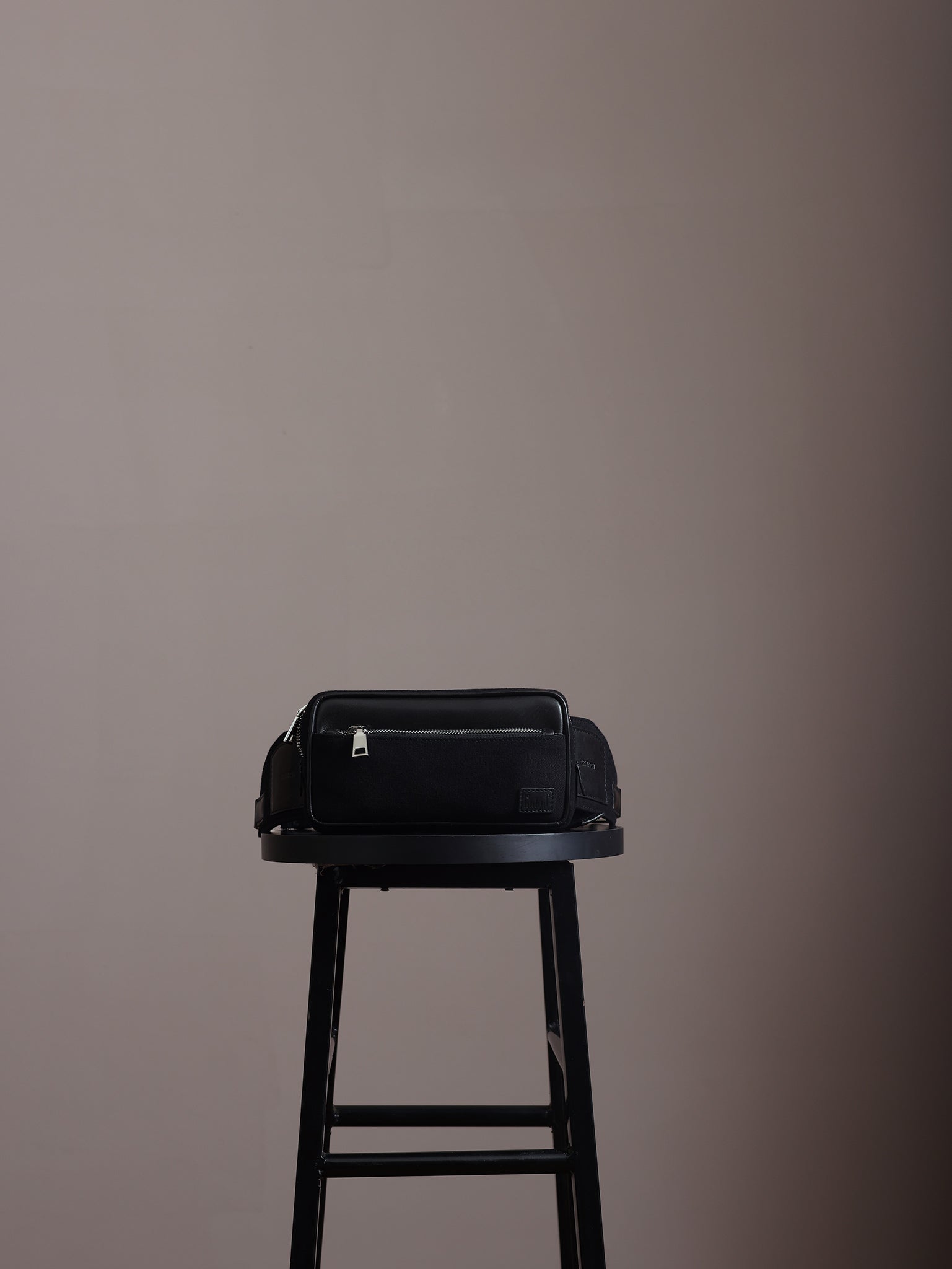 Minimal studio shot of the Studio Ek Inch Metropolitan crossbody bag in black waterproof canvas and reclaimed leather placed on a stool, featuring a front zip pocket and premium hardware. Designed as a premium crossbody bag for men, it blends contemporary aesthetics with functional storage for travel and everyday use.