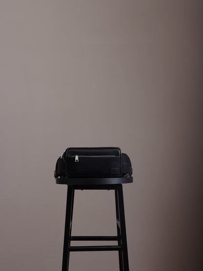Minimal studio shot of the Studio Ek Inch Metropolitan crossbody bag in black waterproof canvas and reclaimed leather placed on a stool, featuring a front zip pocket and premium hardware. Designed as a premium crossbody bag for men, it blends contemporary aesthetics with functional storage for travel and everyday use.